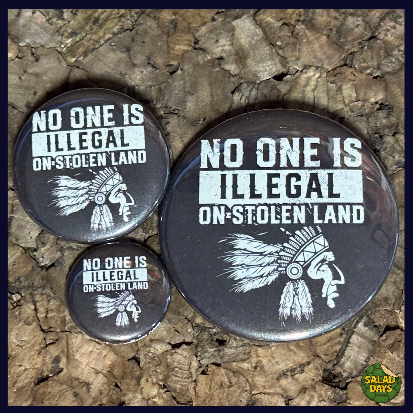 Three buttons with 'No One Is Illegal On-Stolen Land' text and native American headdress graphics on a textured surface.
